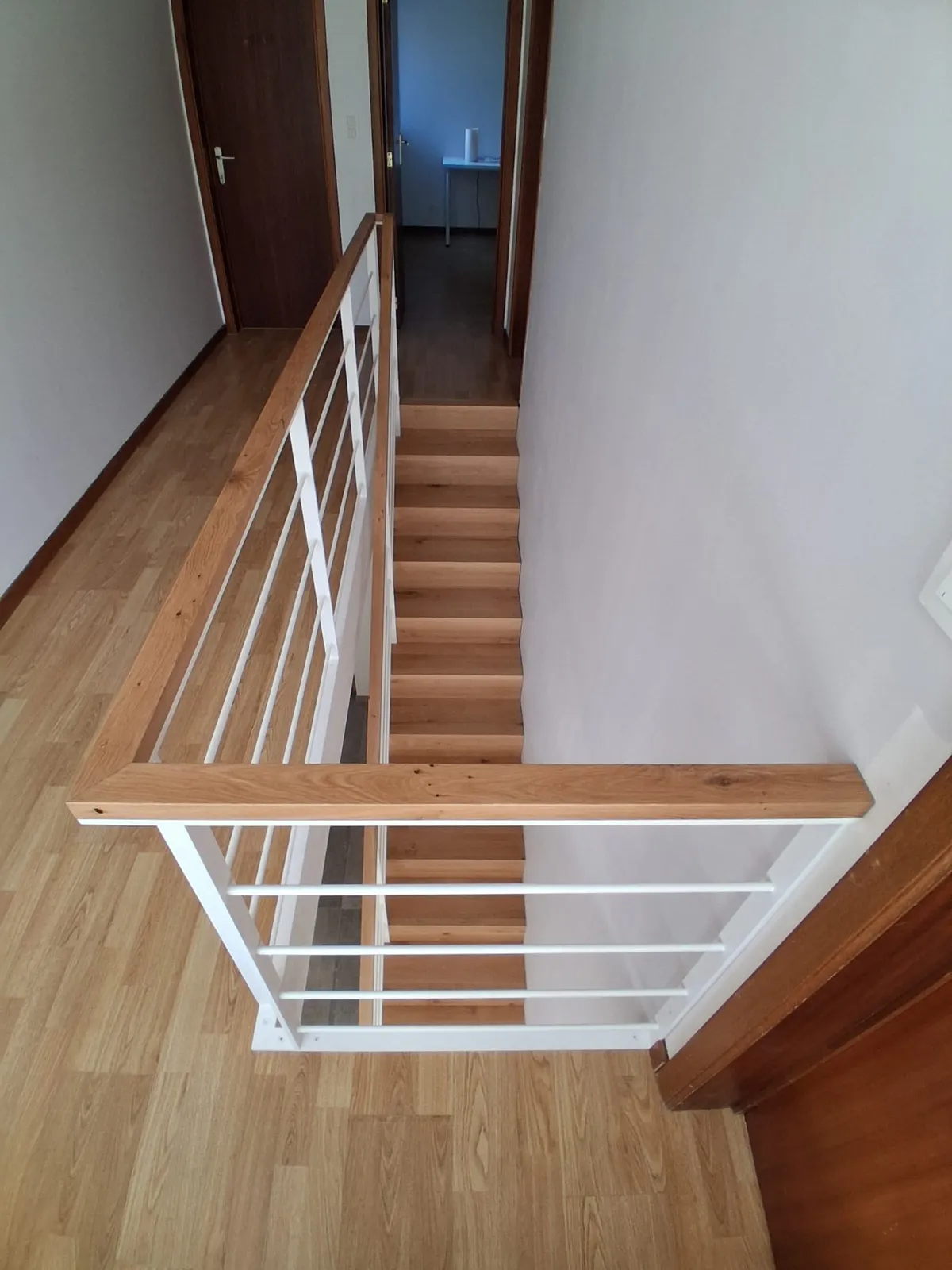 Escalier en bois vu de l'étage, derrière un garde-corps blanc à main courante en bois